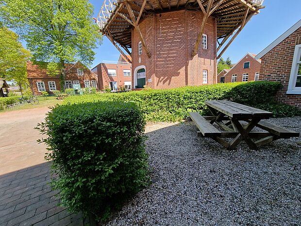 Rastplatz an der Hahnentanger Mühle, Gemeinde Rhauderfehn, Ostfriesland Holztisch mit Bänken auf Kiesfläche neben grünen Hecken vor einer roten Backsteinmühle und weiteren Backsteingebäuden unter blauem Himmel
