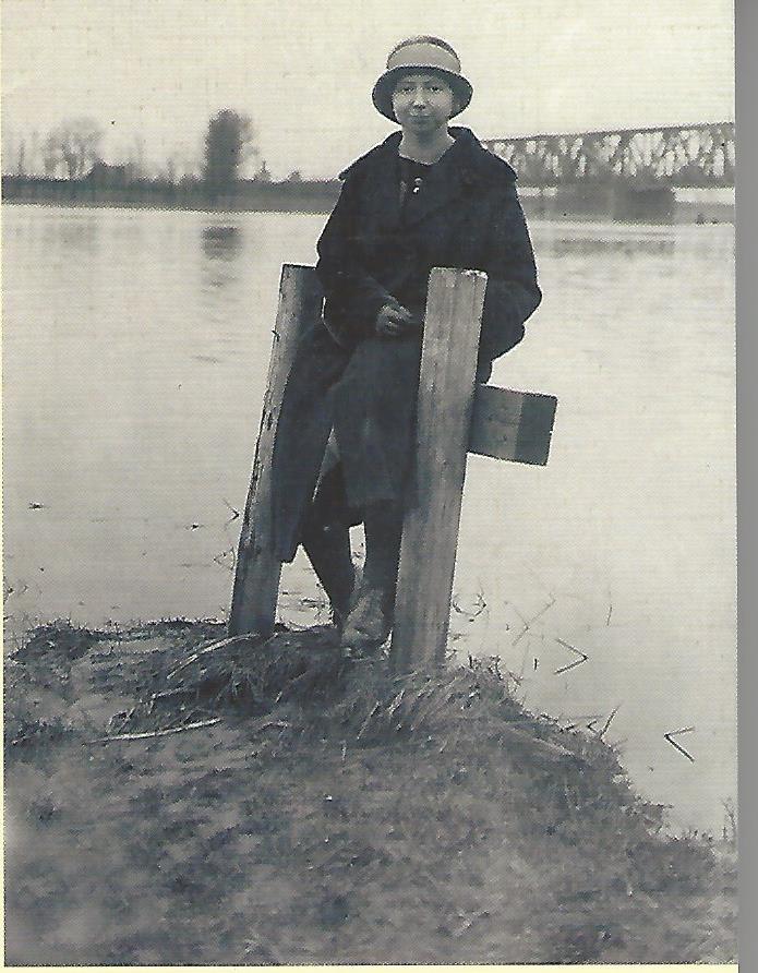 Menna Steen sitzt auf einem Holzpfahl am Wasser, im Hintergrund eine Brücke.