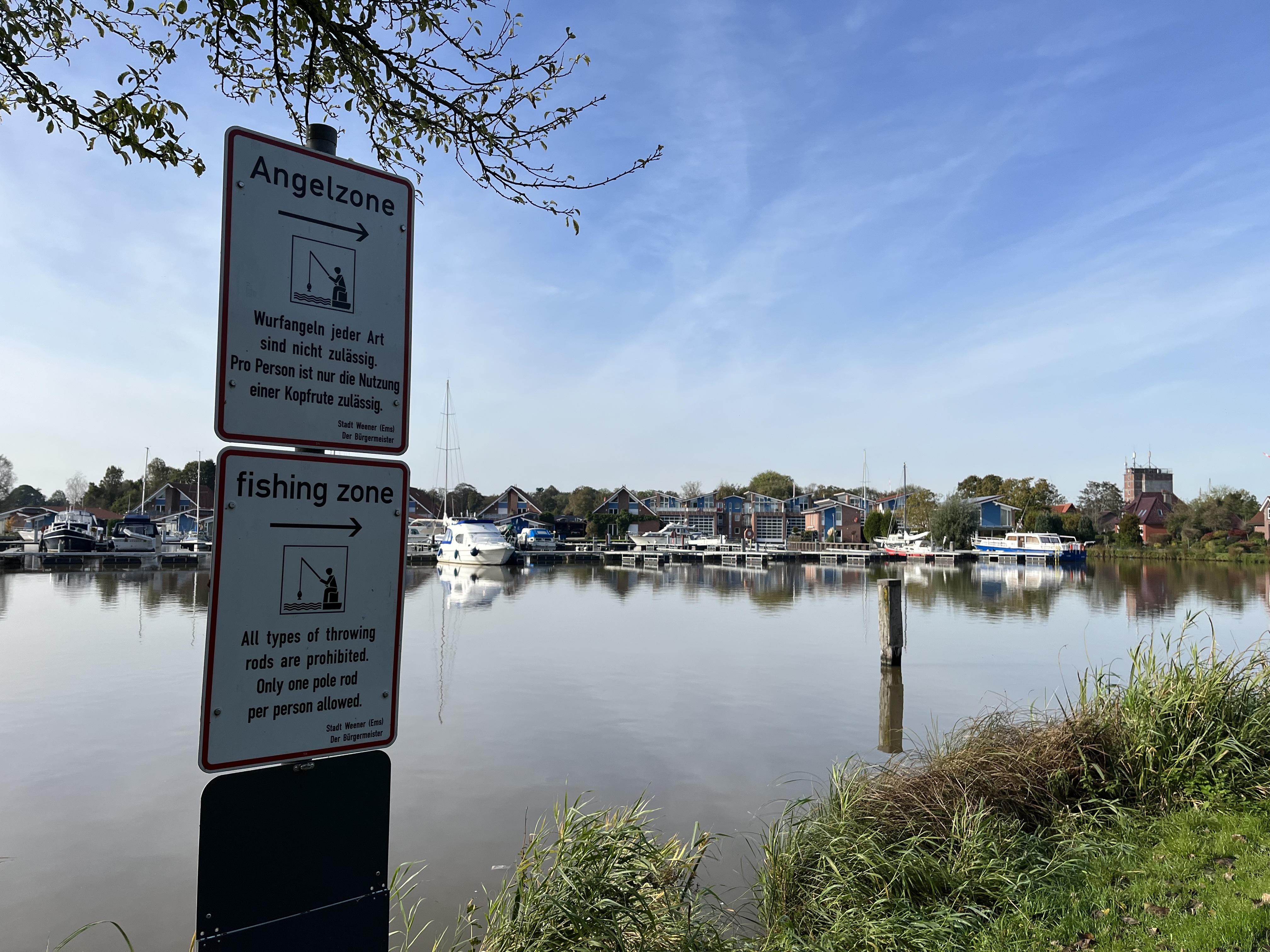 Foto: Blick auf die anliegenden Segelboote. Links im Vordergrund ist ein Schild mit der Beschriftung "Angelzone. Wurfangeln jeder Art sind nicht zulässig. Pro Person ist nur die Nutzung einer Kopfrute zulässig." zu sehen. Darunter ist die Übersetzung angebracht.